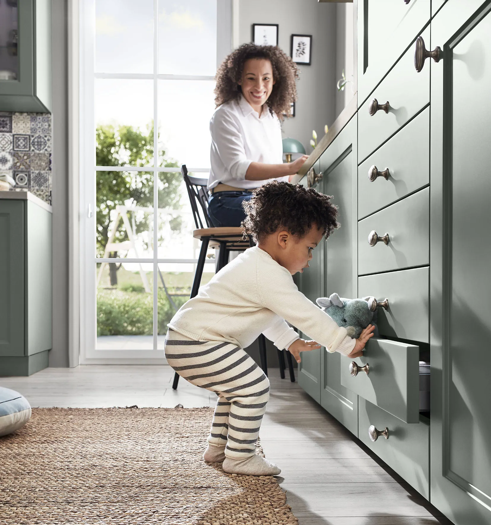 Child playing with a drawer in a sage green kitchen while a mother works at the counter, showcasing a cosy and quiet kitchen atmosphere. Child playing with a drawer in a sage green kitchen while a mother works at the counter, showcasing a cosy and quiet kitchen atmosphere.