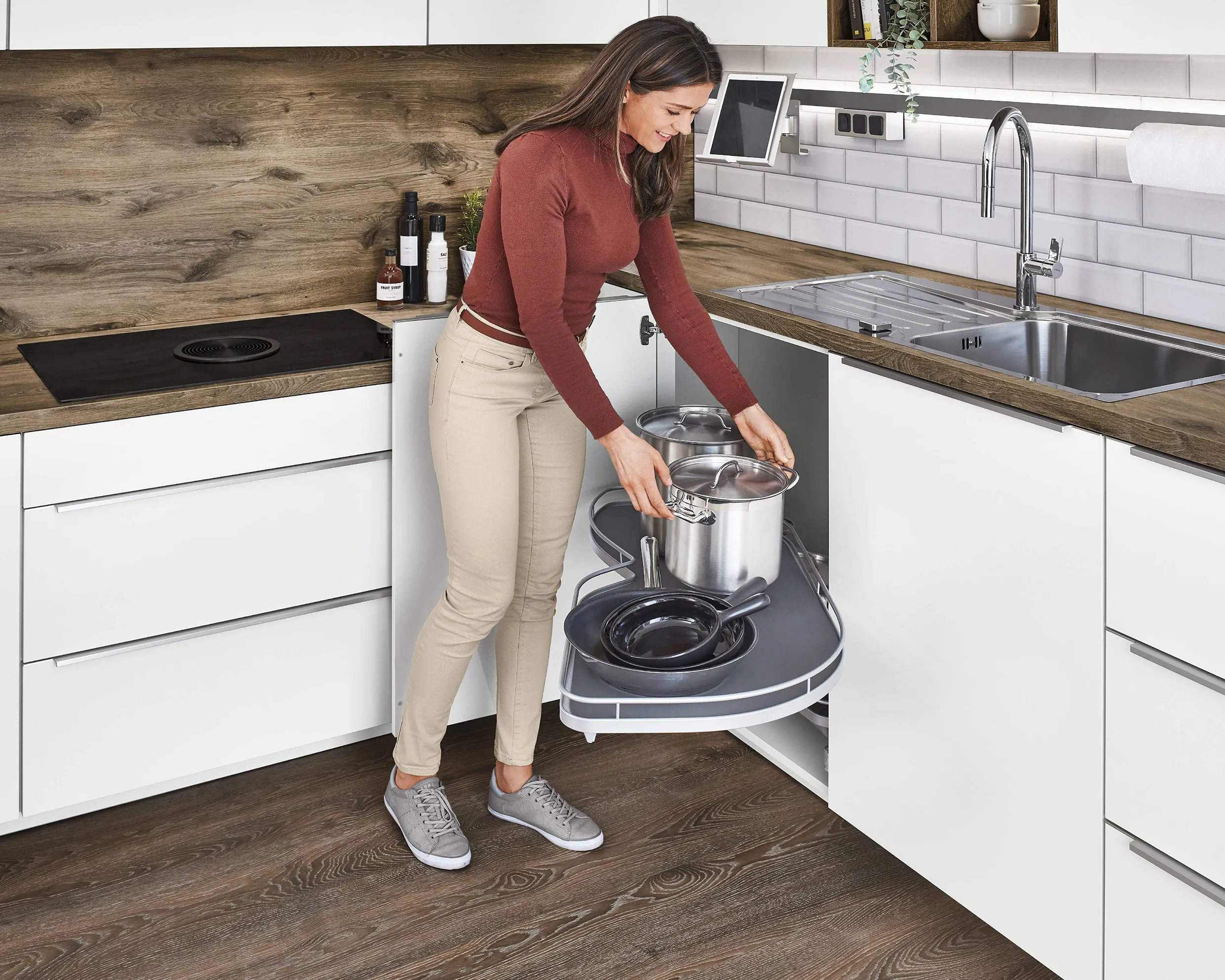 Woman organising pots and pans in a pull-out corner cabinet storage system in a modern kitchen with wooden accents. Woman organising pots and pans in a pull-out corner cabinet storage system in a modern kitchen with wooden accents.
