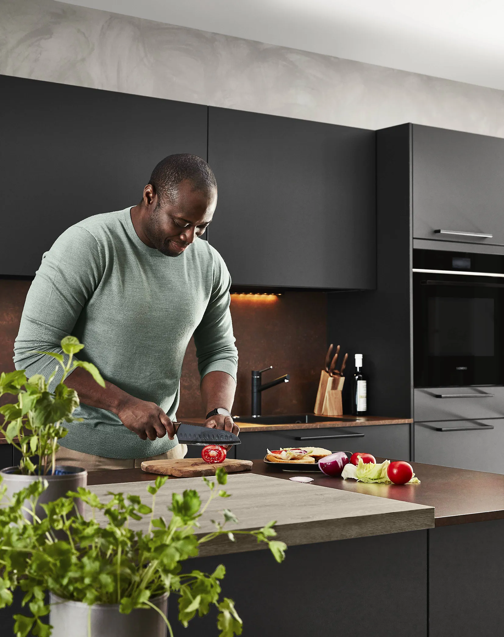 Modern industrial kitchen featuring dark matte cabinets, a concrete-effect backsplash, and a wooden island, with a man preparing fresh vegetables. Modern industrial kitchen featuring dark matte cabinets, a concrete-effect backsplash, and a wooden island, with a man preparing fresh vegetables.