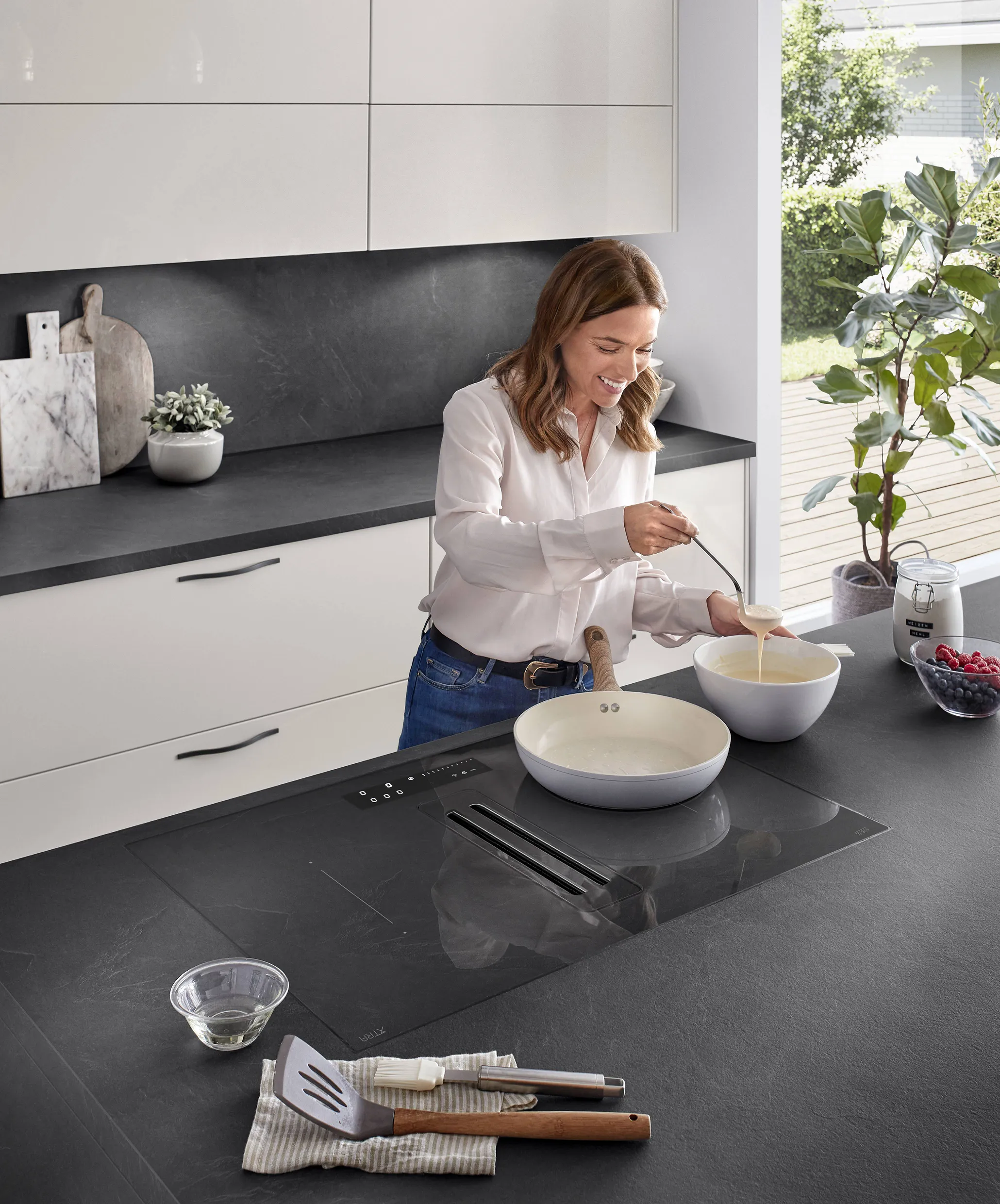 Contemporary kitchen with easy-care black countertops, a sleek induction hob, and modern fixtures, as a woman prepares pancakes near a sunlit window. Contemporary kitchen with easy-care black countertops, a sleek induction hob, and modern fixtures, as a woman prepares pancakes near a sunlit window.