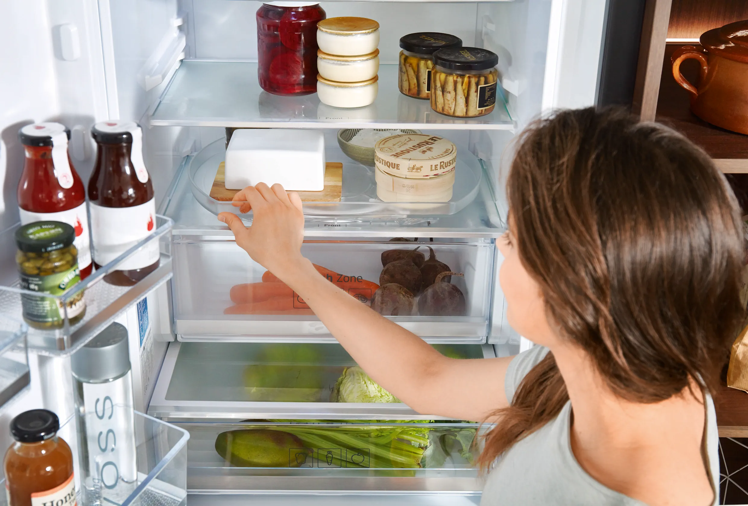 Close-up of a woman organising jars, cheese, and vegetables in a modern fridge, demonstrating efficient storage and accessibility. Close-up of a woman organising jars, cheese, and vegetables in a modern fridge, demonstrating efficient storage and accessibility.