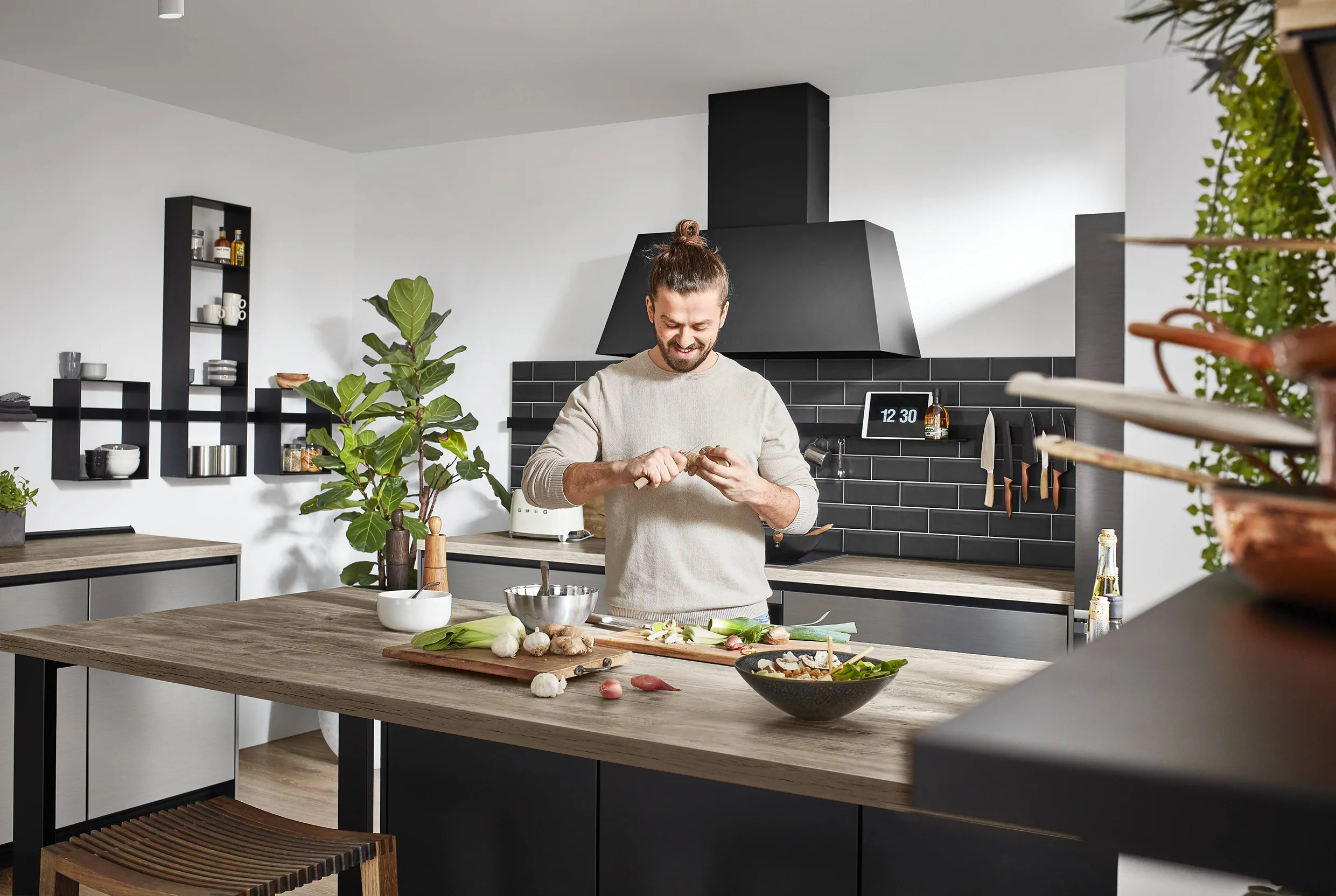 Man preparing ingredients on a natural wood worktop in a modern kitchen with black cabinetry, open shelving, and a stylish black extractor hood. Man preparing ingredients on a natural wood worktop in a modern kitchen with black cabinetry, open shelving, and a stylish black extractor hood.