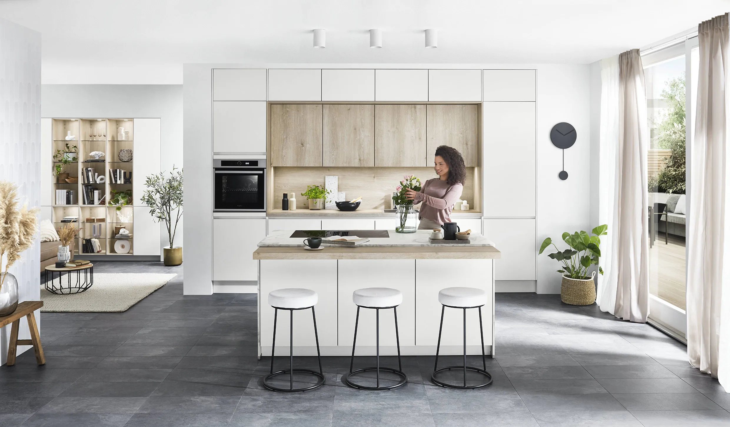 Bright, minimalist kitchen with natural wood accents, featuring open shelves and a woman arranging flowers at the island. Bright, minimalist kitchen with natural wood accents, featuring open shelves and a woman arranging flowers at the island.