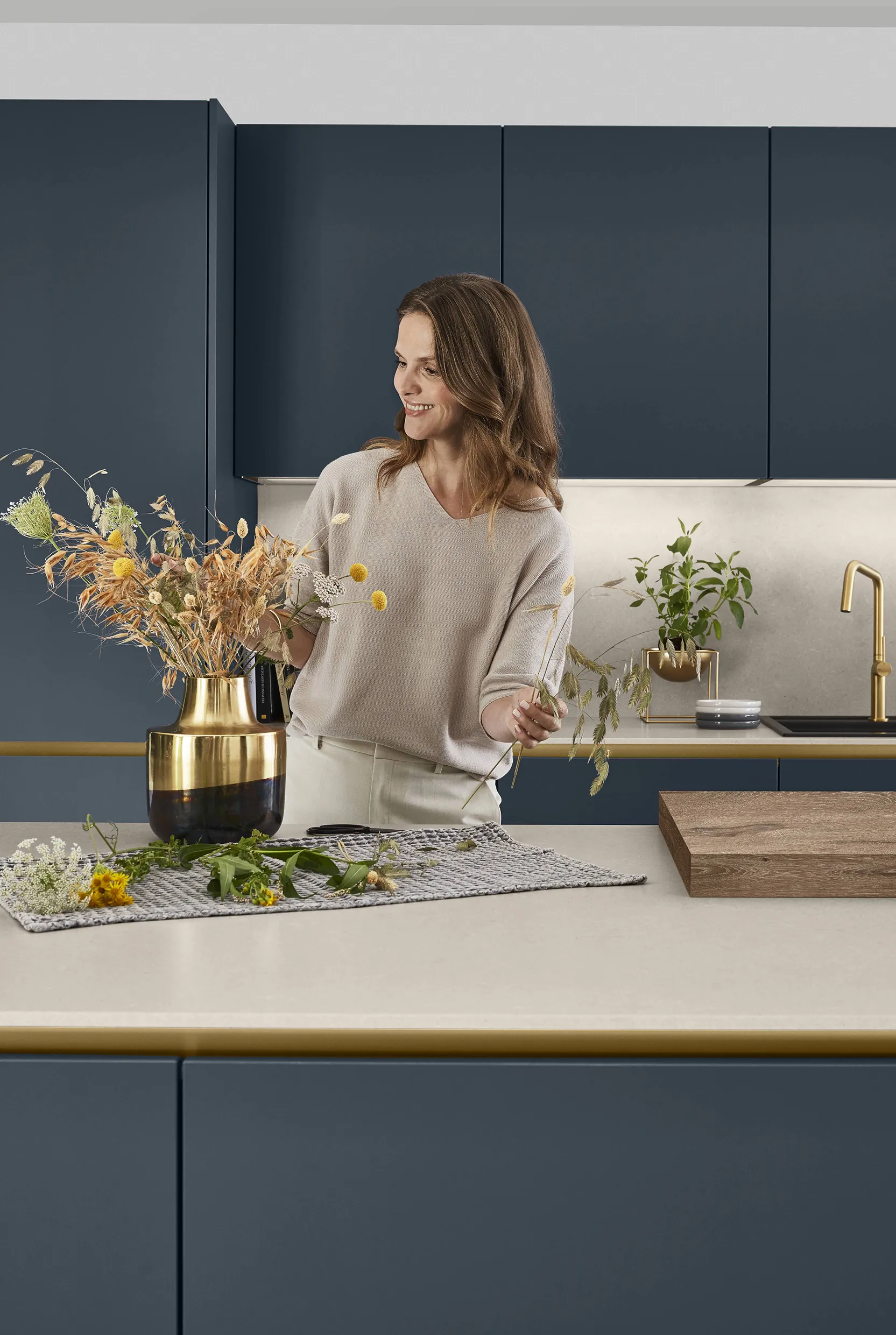 Woman arranging flowers in a stylish kitchen with navy blue cabinets and golden accents, showcasing artful decor and modern design elements. Woman arranging flowers in a stylish kitchen with navy blue cabinets and golden accents, showcasing artful decor and modern design elements.