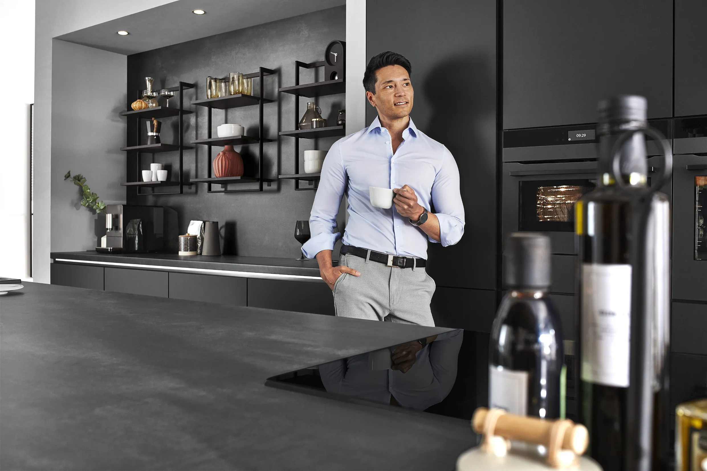 Man enjoying a coffee in a sleek black kitchen with metallic appliances and open shelving, showcasing a modern industrial design. Man enjoying a coffee in a sleek black kitchen with metallic appliances and open shelving, showcasing a modern industrial design.