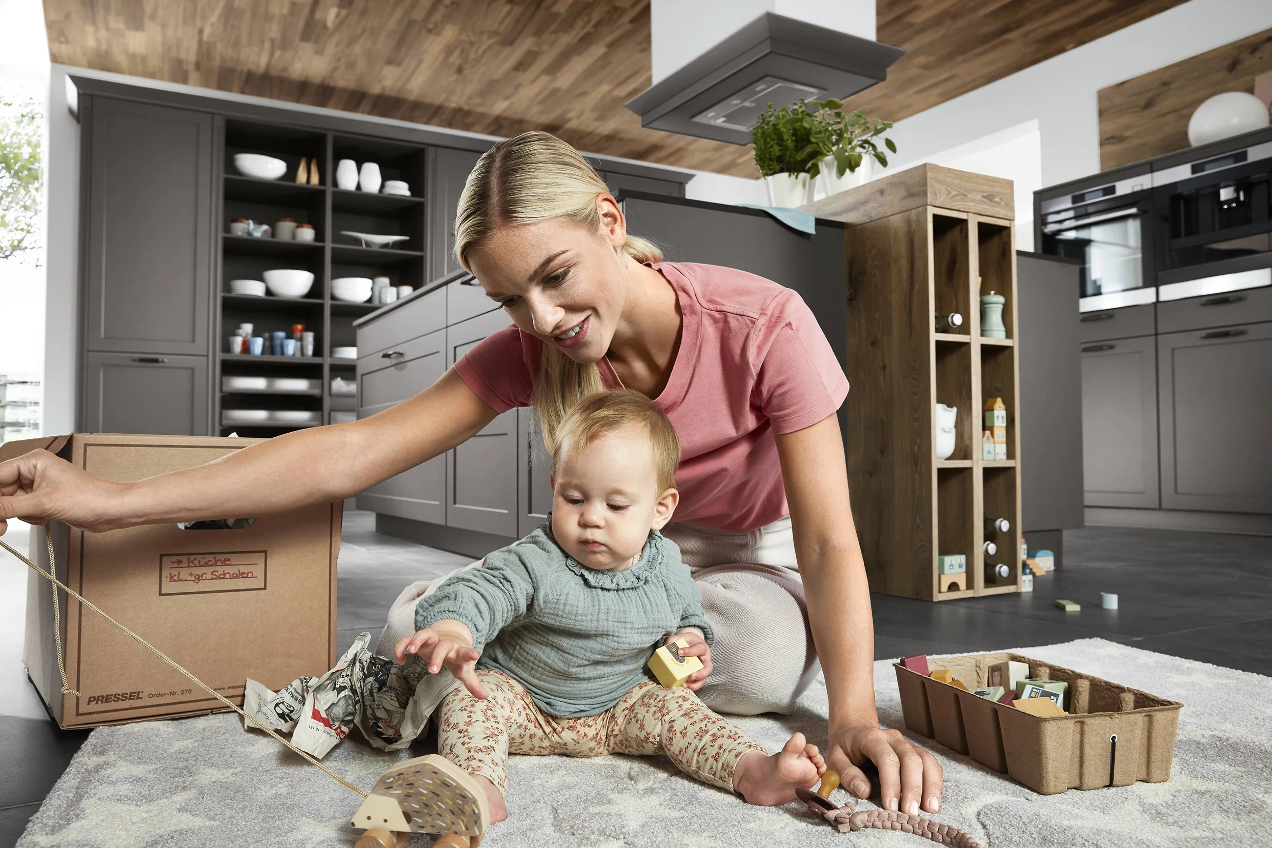 Mother and child unpacking in a grey kitchen with a wooden ceiling and dark tile flooring, highlighting a functional and family-friendly design. Mother and child unpacking in a grey kitchen with a wooden ceiling and dark tile flooring, highlighting a functional and family-friendly design.