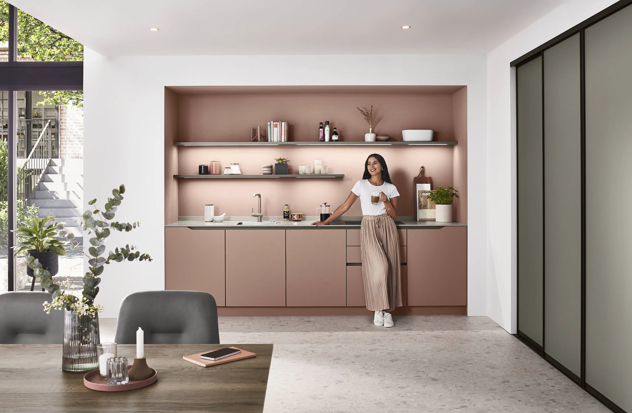 Woman enjoying coffee in a minimalist pink kitchen with open shelving and streamlined cabinets, embodying a modern and simplified design. Woman enjoying coffee in a minimalist pink kitchen with open shelving and streamlined cabinets, embodying a modern and simplified design.