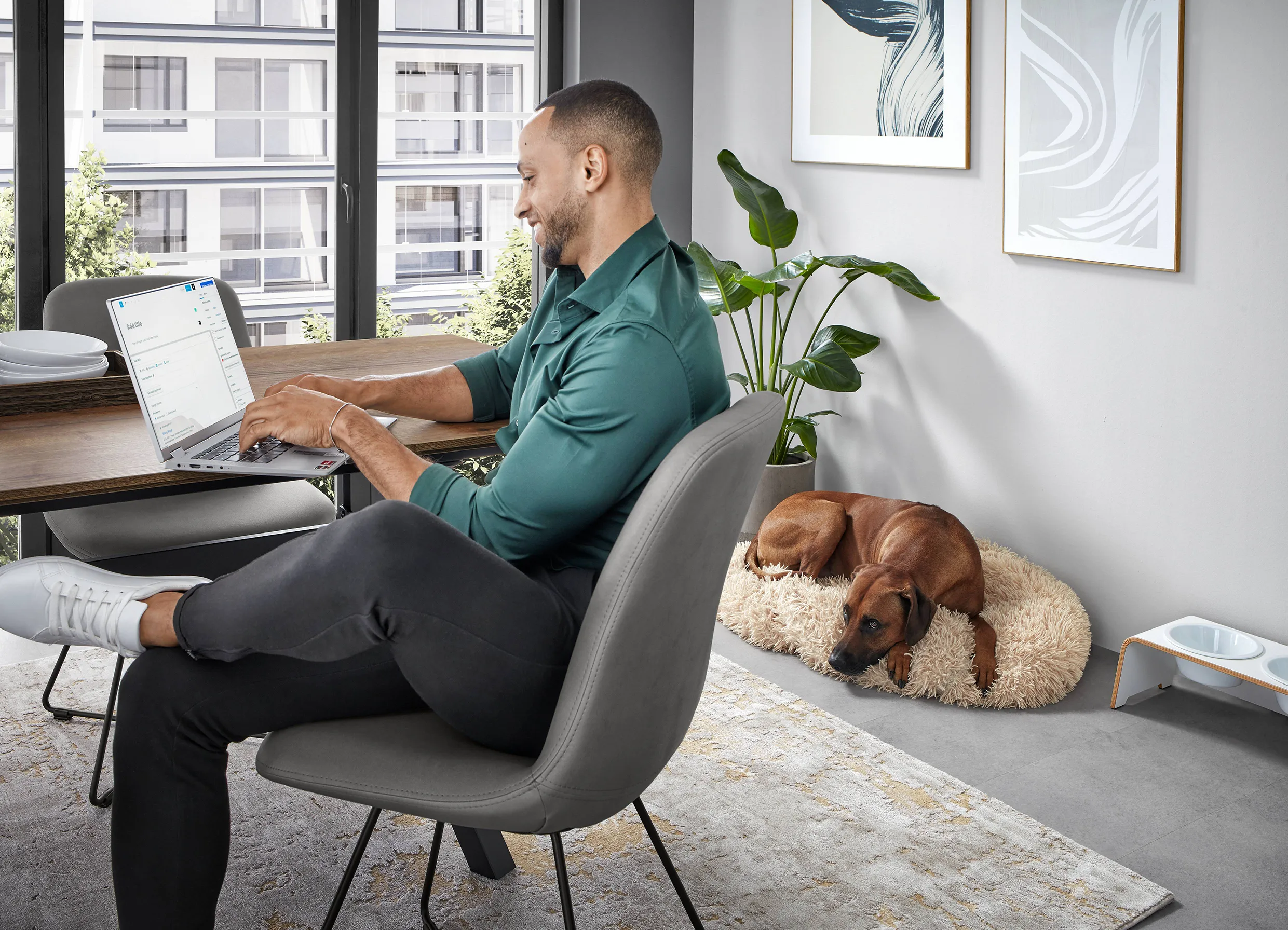 Man working on a laptop in a bright, modern home with natural light, a dog resting on a plush bed beside him. Man working on a laptop in a bright, modern home with natural light, a dog resting on a plush bed beside him.