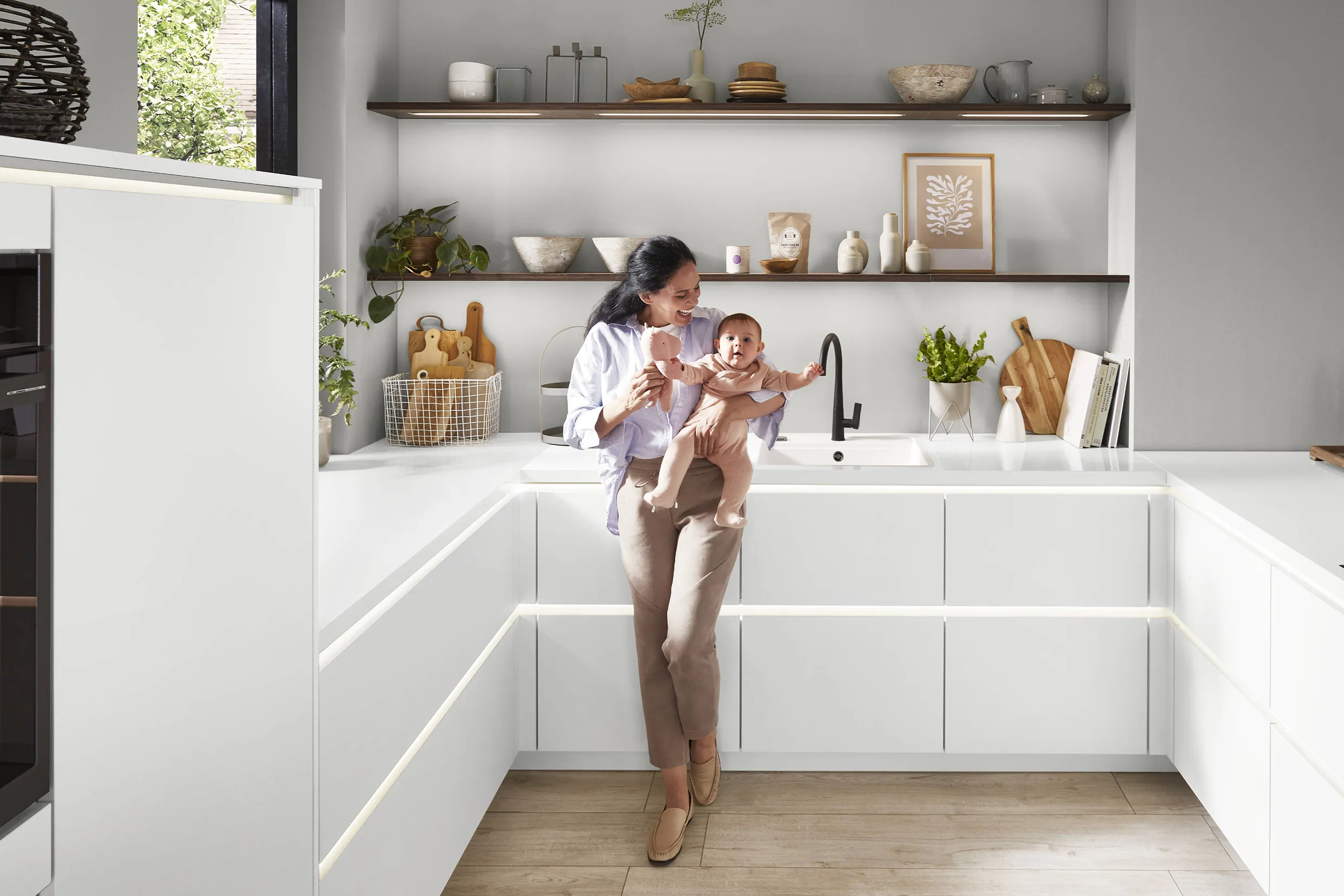 Mother holding her baby in a modern kitchen with white minimalist cabinets, open shelving, and a black matte tap, showcasing a blend of functionality and warm aesthetics. Mother holding her baby in a modern kitchen with white minimalist cabinets, open shelving, and a black matte tap, showcasing a blend of functionality and warm aesthetics.
