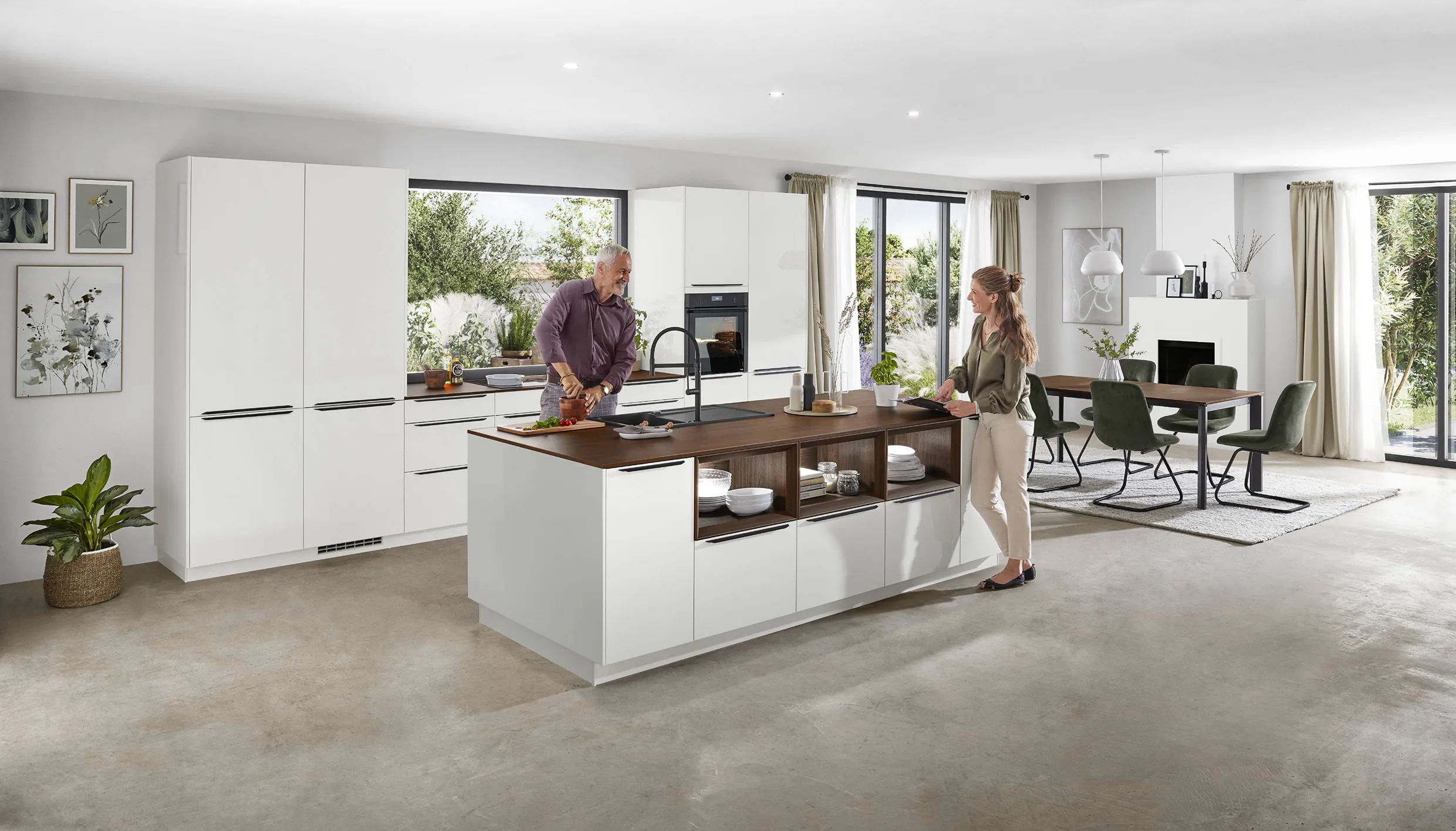 Couple preparing food in a spacious white kitchen with a large island, open shelving, and natural light, showcasing modern design with wooden accents. Couple preparing food in a spacious white kitchen with a large island, open shelving, and natural light, showcasing modern design with wooden accents.